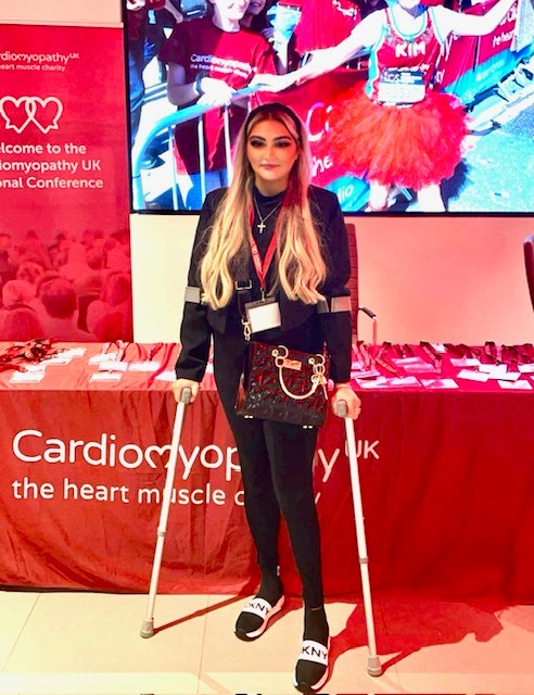 Charlie, a young woman with long blonde hair, stands with crutches in front of a Cardiomyopathy UK table display.