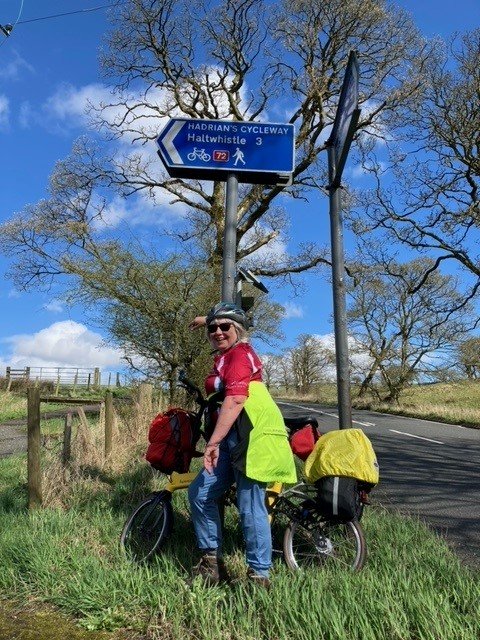 Sue stands with her bicycle under a road sign pointing towards Hadrian's wall.